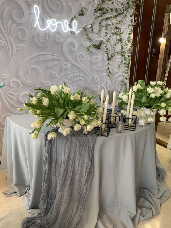 Оформление стола молодоженов для экспозиции на свадебной выставке. Неоновая вывеска, композиция из искусственных цветов. Фон за молодоженами